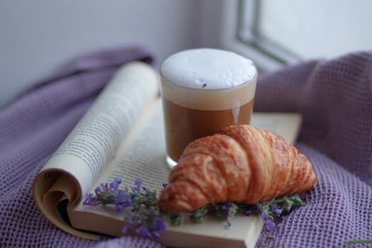 NK#03: Học trò và những gói túi mù A delightful breakfast scene featuring a croissant, cappuccino, and an open book with lavender accents.